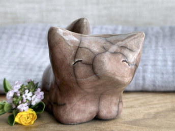 Sitting raku ceramic kitsune fox sculpture glazed in warm brown with hints of mauve. It has a very kind, gentle face, and its tail is curled up its back. It has two little feet at the front and rather large, pointy ears.