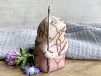 raku ceramic nature spirit warrior sculpture. it is glazed in a bottom-to-top gradient of pale soft red to pale peach and carries a brown stick in one of its arms. it has a sad and worried but determined look on its gentle face.