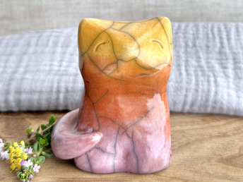 Raku ceramic sitting fox glazed in a gradient from soft mauve to orange to golden yellow (bottom to top). It has a kind face, and its tail is curled around its body on its right side.