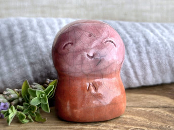 a smiley and loving Jizo Shinto raku ceramic sculpture talisman glazed in a bottom-to-top gradient of warm red-orange to a soft pinky red. they have a kind, gentle face and are a wonderful smooth shape to hold in the hand.