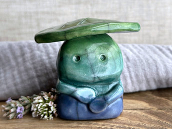 Raku ceramic sculpture glazed in a bottom-to-top gradient of blue to forest green to leaf green with a kind, but slightly intense and worried, face, and a leaf-green leaf on its head. It also has two cute little arms.