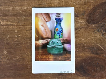 an instax mini print of a small, forest green raku ceramic kodama with a gentle face and two cute little arms. it's on a wooden shelf, and there are various pieces of wood and also a small bottle painted with plant designs in blue, green, purple and orange behind it.