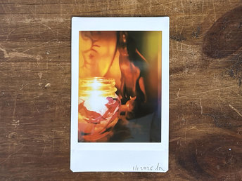 a dark, atmospheric instax mini print of a blue raku ceramic kodama guardian nature spirit with a white crescent moon on their head. the scene is lit by a candle in a glass jar that's covered in tissue paper in autumnal reds, oranges, golden yellows, and purples. there's a wooden hare in the background along with the edge of a traditional drum.