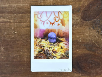 an instax mini print of a small, violet guardian nature spirit. there are lots of dried, pale golden orange calendula flowers and lavender flowers around them. behind them is a stick wrapped in plant-dyed wool in warm pink and orange, and behind that is the bottom of a traditional drum decorated with henna.