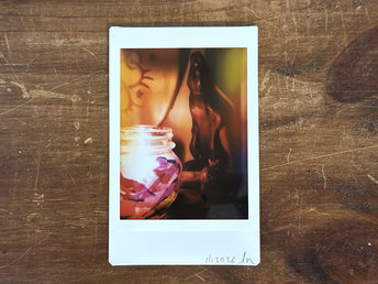 a dark, atmospheric instax mini print of a blue raku ceramic kodama guardian nature spirit with a white crescent moon on their head. the scene is lit by a candle in a glass jar that's covered in tissue paper in autumnal reds, oranges, golden yellows, and purples. there's a wooden hare in the background along with the edge of a traditional drum.