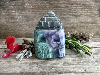 Tiny raku ceramic cottage glazed in soft forest green, blue, indigo, and violet-purple. Its roof is glazed in grey and carved to look like slates. It has a carved door and windows and lots of plant and flower carvings too.
