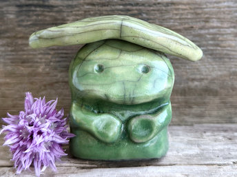 Raku ceramic sculpture glazed in forest green to spring green (bottom to top) with a sweet face and a lighter, yellow-green leaf on its head. It also has two cute little arms.