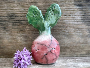 a very smiley and happy radish spirit raku ceramic sculpture. it has a round body glazed in a soft radish red colour. the top of its body fades to white, and it has two green leaves on the top of its head. body. it is round with green leaves on top!