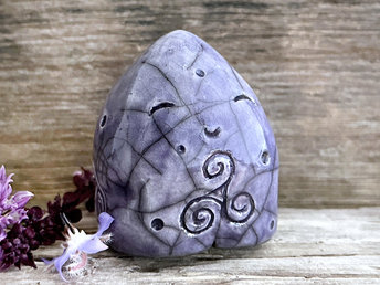 round and vaguely egg-shaped nature spirit sculpture. it is glazed in violet and has spiral carvings, including a triskelion, on its body. it also has a gentle, happy face.