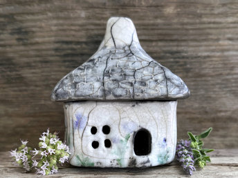Rustic raku ceramic kurinuki cottage glazed in white with pale green plant and pale violet flower patterns on the walls. The roof has a chimney and is glazed in grey with hints of white to look like slate. It has a cute little doorway and lots of windows.
