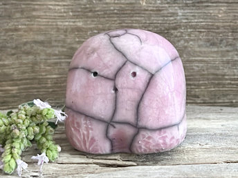 a small and stout nature spirit raku sculpture glazed in pale pink-mauve. it has a cute little face, and little plant carvings on its front.