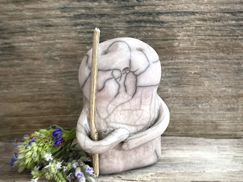 raku ceramic nature spirit warrior sculpture. it is glazed in very pale, soft mauve colour and carries a brown stick in one of its arms. it has a sad and worried but determined look on its gentle face.