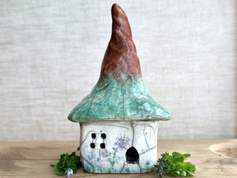 a rustic raku ceramic kurinuki cottage glazed in white with green plant carvings and purple, violet, and indigo flower carvings on the walls. the roof has a very tall, pointy and slightly twisty chimney and is glazed in brown on the pointy bit and shades of green on the lower bit to look a bit like a tree. the cottage has a cute little doorway and lots of windows.