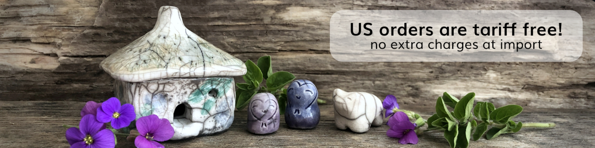 against a background of driftwood, a rustic, raku ceramic cottage glazed in white with green leaves and purple flowers on its walls. with it are two jizo sculptures — one lavender in colour and the other blue — and a white raku bear. there are purple flowers and green leaves on the wood around them.